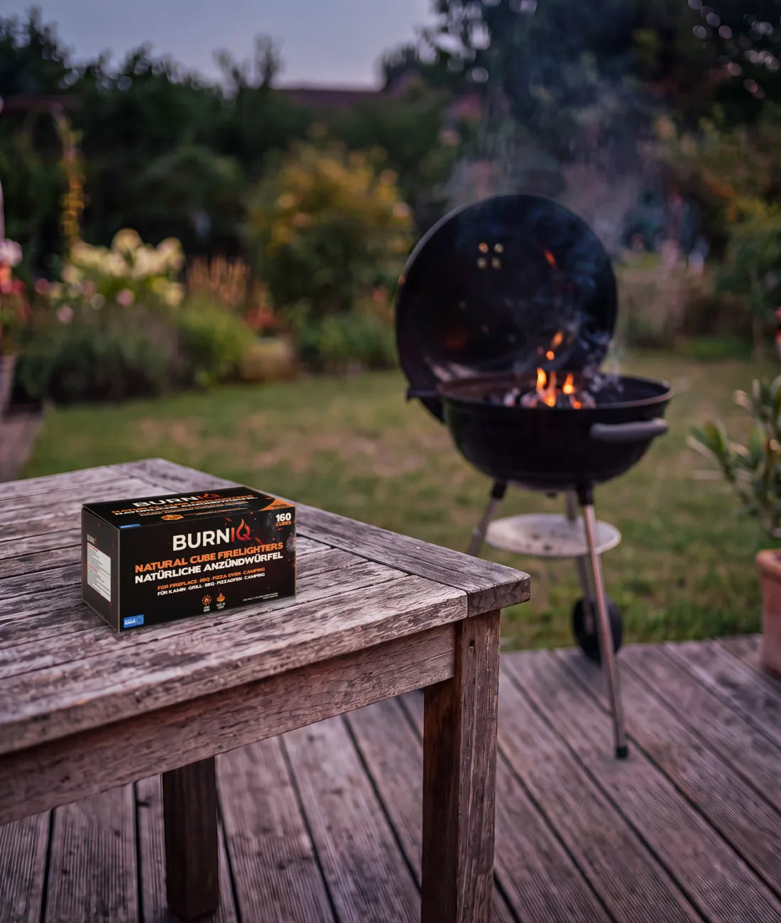 BURNIQ Anzündwürfel-Box auf einem Gartentisch, dahinter ein Kugelgrill mit glühender Holzkohle