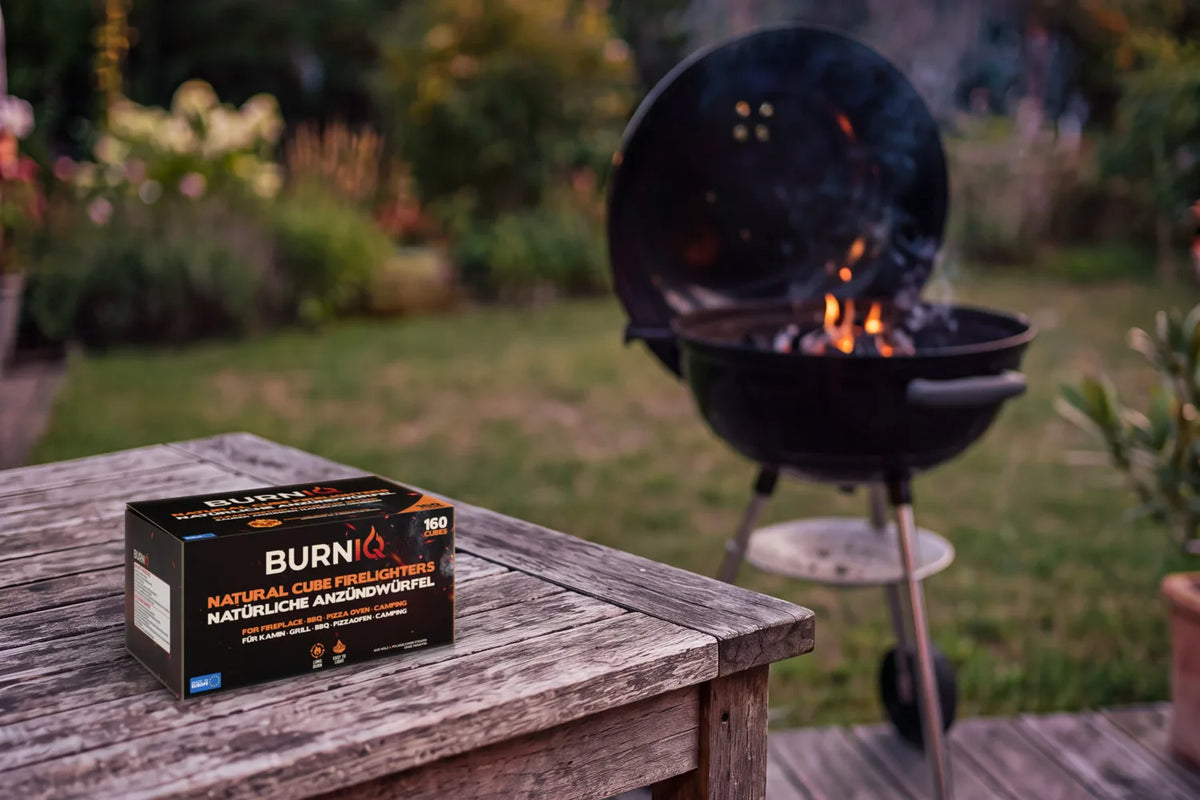 BURNIQ Anzündwürfel-Box auf einem Gartentisch, dahinter ein Holzkohlegrill